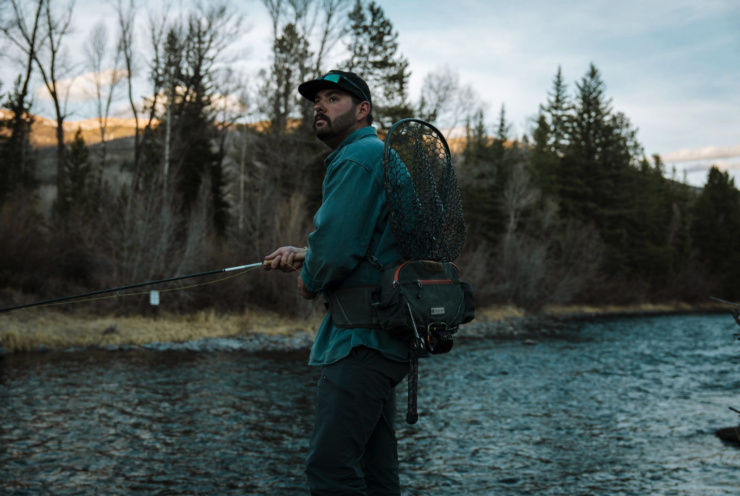 Angler holding fly rod wearing Fishpond Elkhorn Lumbar Pack Duskwood Waxed Cotton