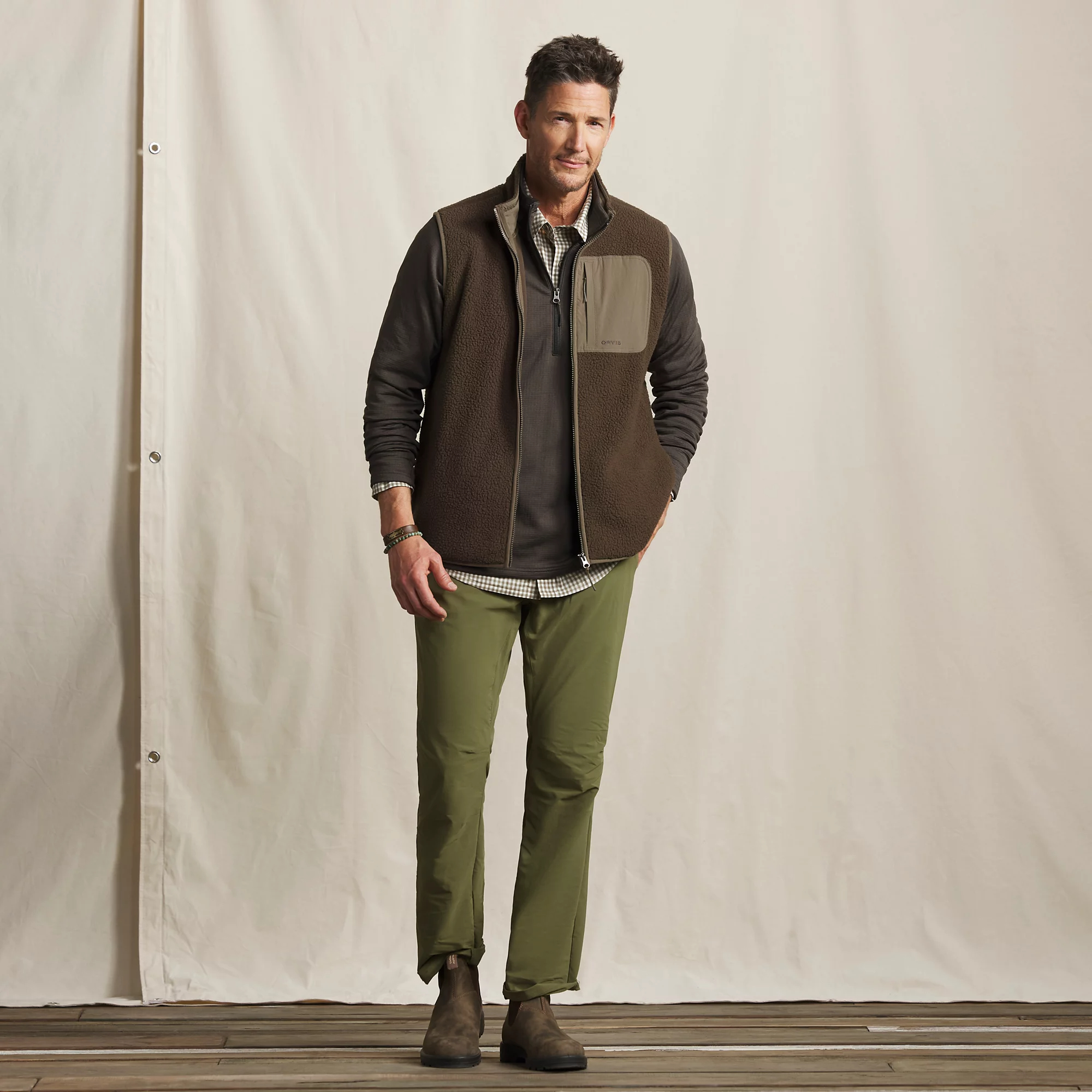 Full-length view of man wearing brown Orvis Mad River fleece vest over layered shirts with green pants for casual outdoor wear.