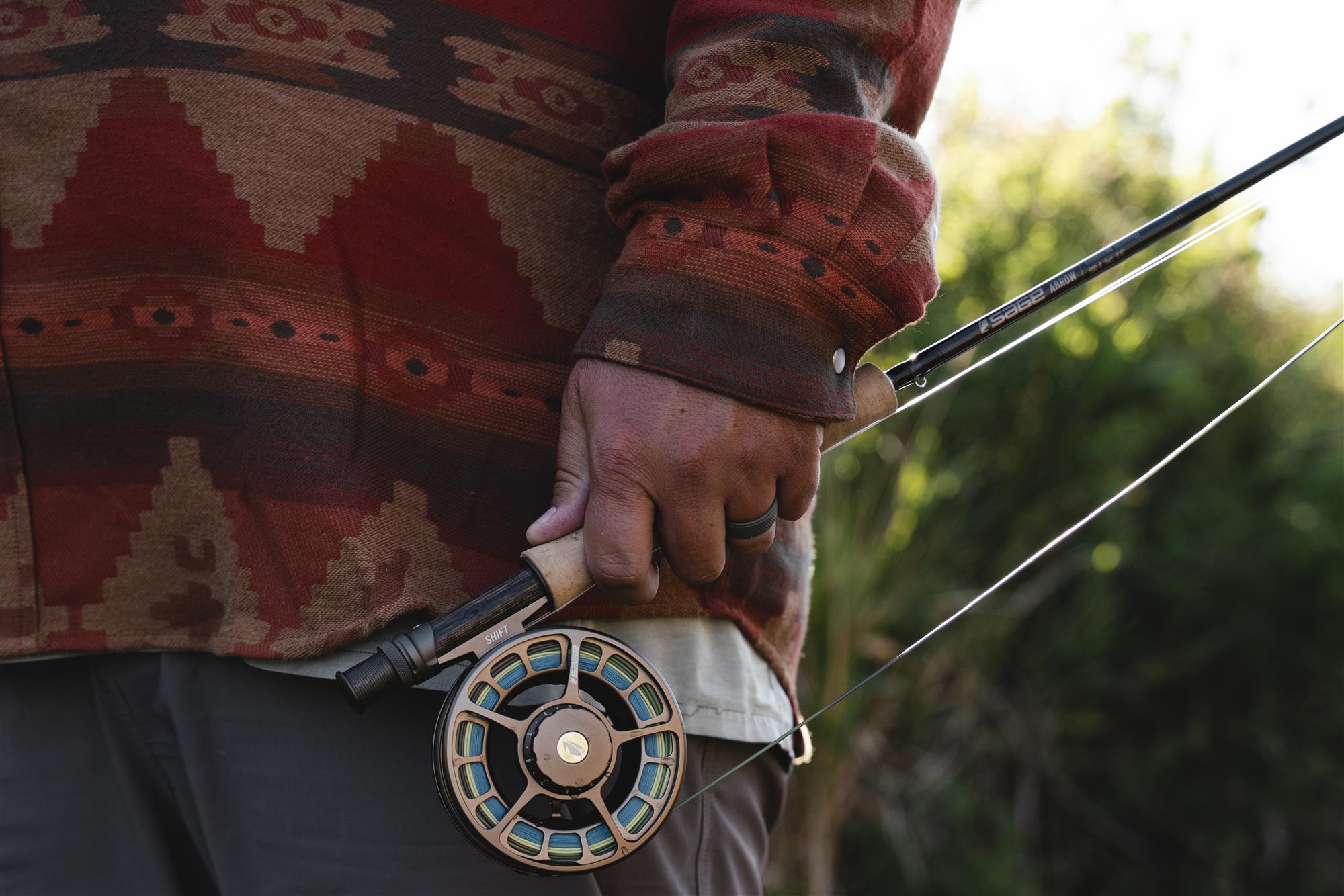 Angler holding a fly rod with a Sage SHIFT fly fishing reel at their side—outdoor fly fishing lifestyle shot near the water