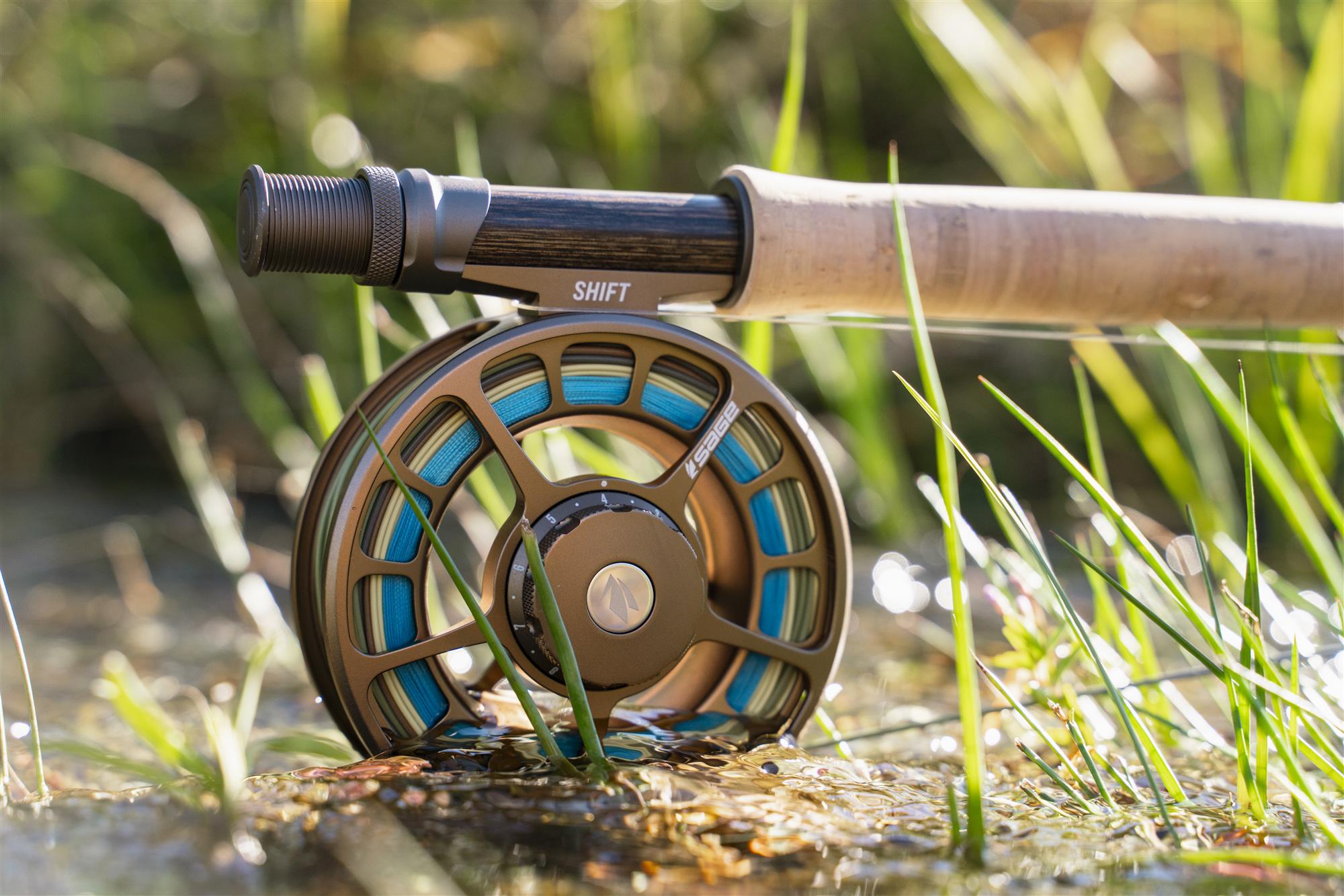 Sage SHIFT fly fishing reel on a cork-handled fly rod resting in shallow creek water with riverside grass (