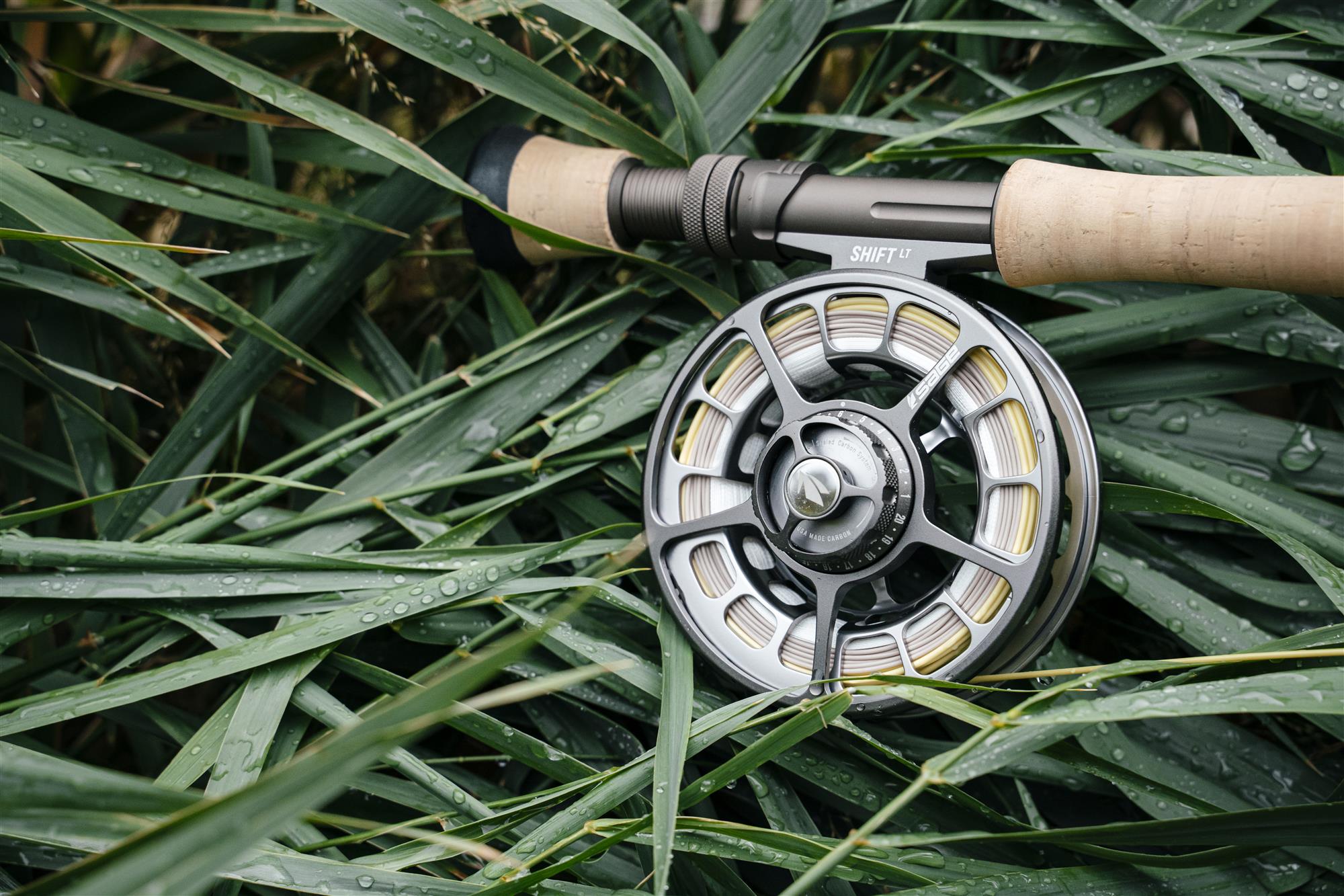 Sage SHIFT LT fly fishing reel mounted on a cork-handled fly rod resting in wet grass—durable anodized reel detail photo