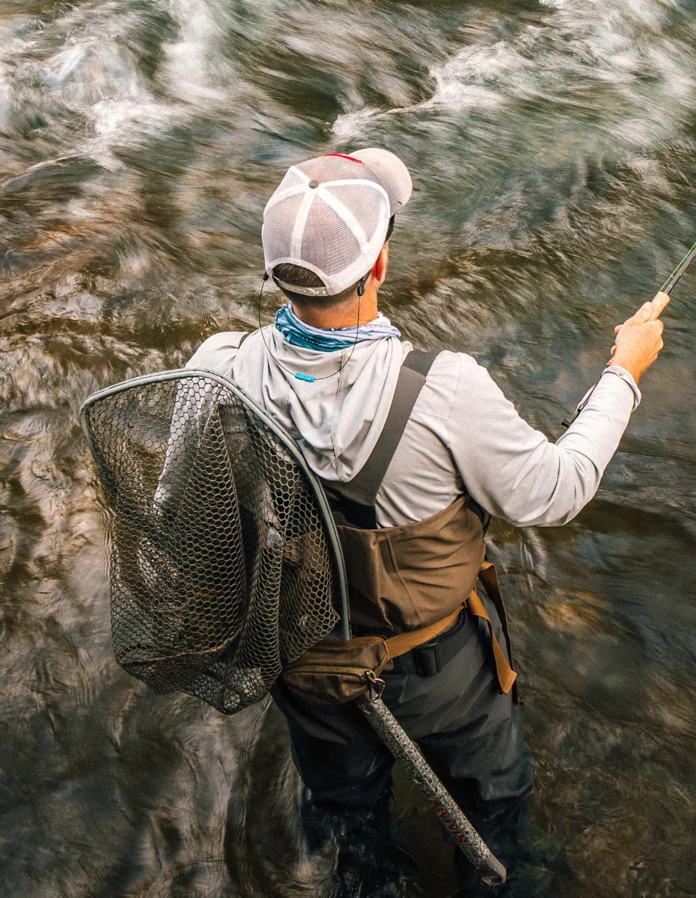 Fly angler wearing Simms G3Z Pro Guide waders while casting in a flowing river with a landing net