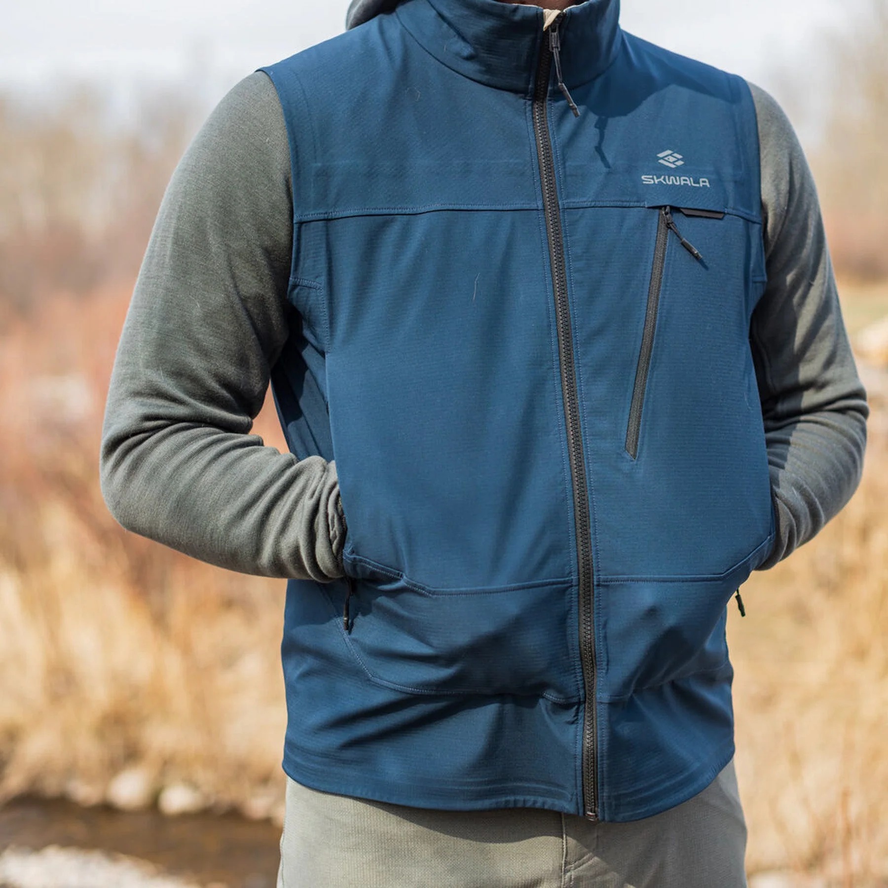 Blue Skwala Backeddy fishing vest layered for cool-weather fly fishing with zippered handwarmer pockets