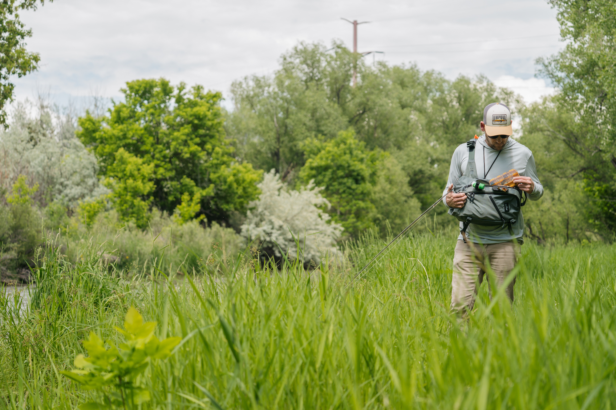 Fly fisherman accessing gear from Umpqua sling pack in tall grass riverbank fishing modular sling pack setup